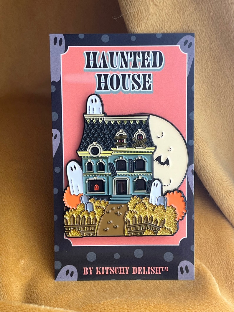 May include: A black and white enamel pin featuring a haunted house with a green door, boarded windows, and a moon with a ghost face. The house is surrounded by a fence and bushes. The pin says "Haunted House" and "By Kitschy Delish&trade;".