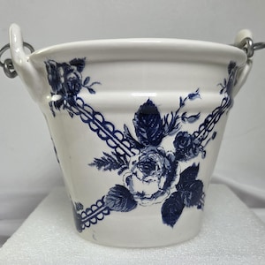 May include: A white ceramic bucket featuring a dark blue floral pattern with roses and decorative lattice trim. The bucket has two integrated side handles with attached metal rings for hanging, providing a functional and classic design element.