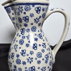 May include: A cream-colored ceramic pitcher with a matching lid, featuring a repeating pattern of small blue flowers, dots, and swirls. The pitcher has a curved handle, a wide spout, and a glossy finish, showcasing a hand-painted aesthetic.