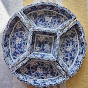 May include: A ceramic serving set consisting of four fan-shaped dishes arranged around a central square bowl. Each piece features a dark blue background with a repeating pattern of white and light orange flowers, green stems, and swirling lines.