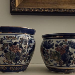 May include: Two round ceramic planters featuring a white base with blue, pink, and green floral patterns accented by gold outlines. Both pots have a solid dark blue rim and base, with a thin gold band bordering the top and bottom edges.