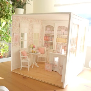 K&ouml;nnte beinhalten: Ein Miniatur-Puppenhaus-Ladeninterieur mit hellem Holzboden, gebl&uuml;mter Tapete und wei&szlig;en M&ouml;beln. Der Raum enth&auml;lt einen kleinen runden Tisch mit zwei St&uuml;hlen, eine Verkaufstheke und hohe Regaleinheiten, die mit kleinen Schachteln und T&uuml;ten gef&uuml;llt sind.