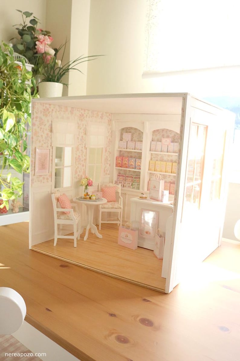 K&ouml;nnte beinhalten: Ein Miniatur-Puppenhaus-Ladeninterieur mit hellem Holzboden, gebl&uuml;mter Tapete und wei&szlig;en M&ouml;beln. Der Raum enth&auml;lt einen kleinen runden Tisch mit zwei St&uuml;hlen, eine Verkaufstheke und hohe Regaleinheiten, die mit kleinen Schachteln und T&uuml;ten gef&uuml;llt sind.