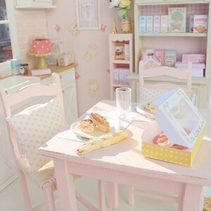 May include: A miniature pink kitchen scene with a table set for a meal. The table has a plate of bread, a glass, and a box of donuts. A pink chair with a polka dot pillow is next to the table. The walls are pink with framed pictures.