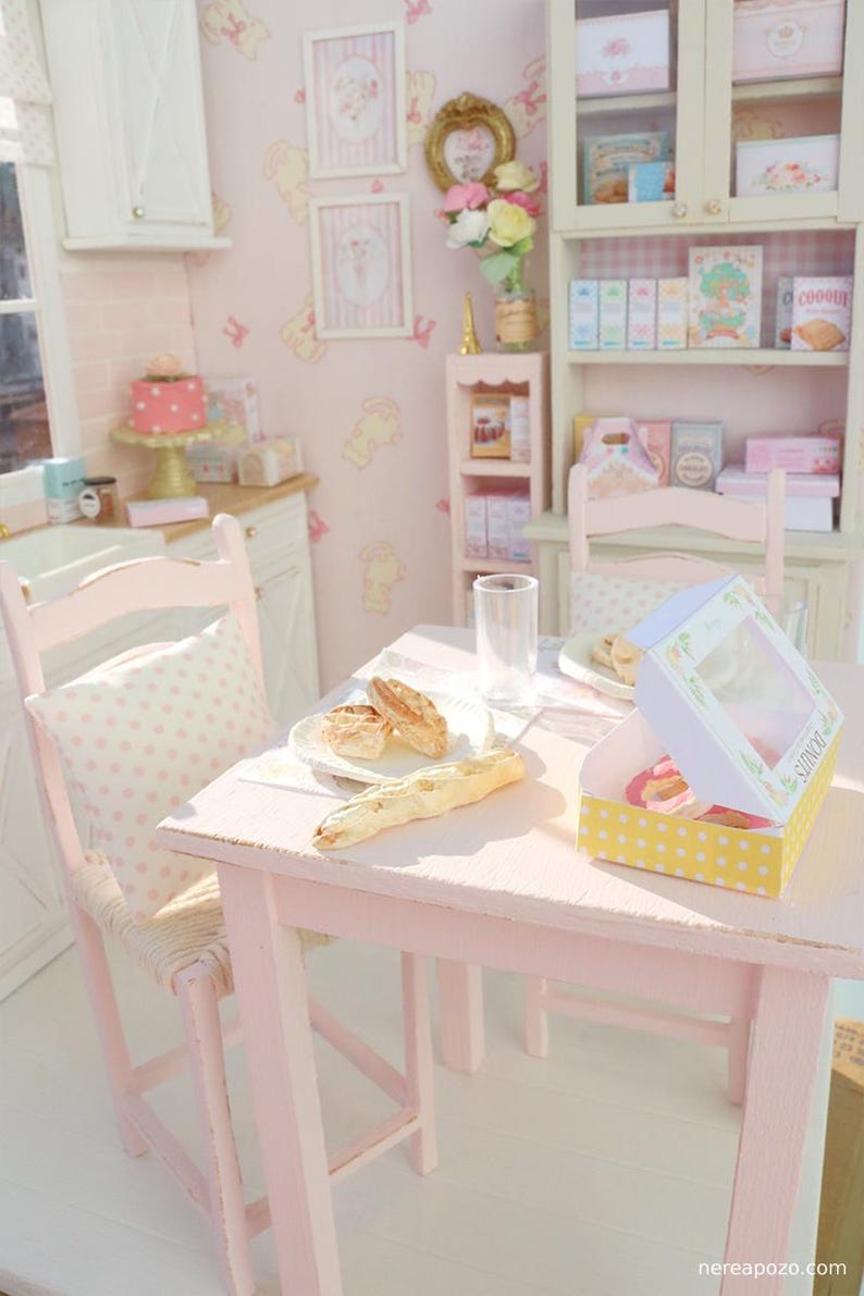 May include: A miniature pink kitchen scene with a table set for a meal. The table has a plate of bread, a glass, and a box of donuts. A pink chair with a polka dot pillow is next to the table. The walls are pink with framed pictures.
