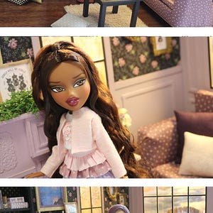 May include: A dollhouse living room scene featuring a doll with long brown hair wearing a pink cardigan and layered skirt, standing near a purple fireplace, a black coffee table, and a purple patterned sofa with various throw pillows.