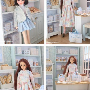 May include: A miniature bakery scene featuring two dolls in a detailed shop setting with shelves stocked with bread, boxes, and pastries. The shop includes a wooden table, stools, and a shopping bag labeled Bakery, all rendered in a light blue and white color palette.