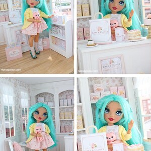 K&ouml;nnte beinhalten: Eine Puppe mit langem t&uuml;rkisfarbenem Haar tr&auml;gt eine gelbe Strickjacke, ein wei&szlig;es T-Shirt und einen rosa gepunkteten Rock. Die Puppe h&auml;lt einen kleinen rosa B&auml;ren. Die Szene zeigt eine Miniatur-B&auml;ckerei mit Regalen, Theke und Cake & Clover-T&uuml;ten.