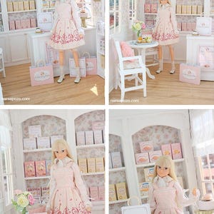 K&ouml;nnte beinhalten: Eine blonde Puppe in einem hellrosa Kleid mit dunkelrosa Blumenmuster, wei&szlig;em Hemd mit Kragen und wei&szlig;en Schuhen, stehend in einer Miniatur-B&auml;ckerei mit wei&szlig;en Regalen, rosa Schachteln und Einkaufst&uuml;ten mit der Aufschrift Cake & Clover.