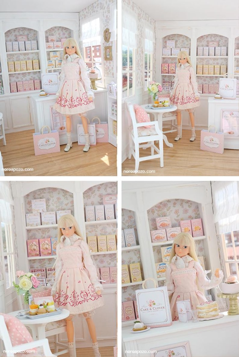 K&ouml;nnte beinhalten: Eine blonde Puppe in einem hellrosa Kleid mit dunkelrosa Blumenmuster, wei&szlig;em Hemd mit Kragen und wei&szlig;en Schuhen, stehend in einer Miniatur-B&auml;ckerei mit wei&szlig;en Regalen, rosa Schachteln und Einkaufst&uuml;ten mit der Aufschrift Cake & Clover.