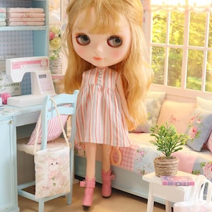K&ouml;nnte beinhalten: Eine Puppe mit blondem Haar, die ein rosa-wei&szlig; gestreiftes Kleid und rosa Stiefel tr&auml;gt. Die Puppe befindet sich in einem Miniaturzimmer mit einer N&auml;hmaschine, einem kleinen Stuhl und einem Bett mit floralen Kissen. Eine kleine Pflanze steht auf einem wei&szlig;en Tisch.