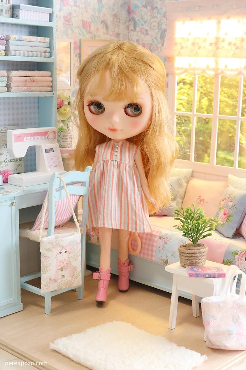K&ouml;nnte beinhalten: Eine Puppe mit blondem Haar, die ein rosa-wei&szlig; gestreiftes Kleid und rosa Stiefel tr&auml;gt. Die Puppe befindet sich in einem Miniaturzimmer mit einer N&auml;hmaschine, einem kleinen Stuhl und einem Bett mit floralen Kissen. Eine kleine Pflanze steht auf einem wei&szlig;en Tisch.