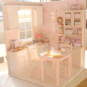 May include: A miniature doll's house kitchen and dining room. The kitchen has white cabinets, a window, and a pink wall. The dining area features a small pink table and chairs. A sign that says "Bakery" is visible.