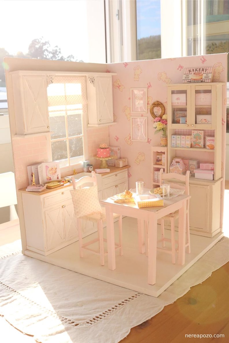 May include: A miniature doll's house kitchen and dining room. The kitchen has white cabinets, a window, and a pink wall. The dining area features a small pink table and chairs. A sign that says "Bakery" is visible.