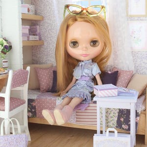 Pode incluir: Uma boneca com longos cabelos loiros e olhos grandes, vestida com um vestido azul claro e botas rosa, com &oacute;culos na cabe&ccedil;a. A boneca est&aacute; sentada em uma cama em miniatura com uma colcha de retalhos. Uma pequena mesa branca cont&eacute;m uma caixa e livros.