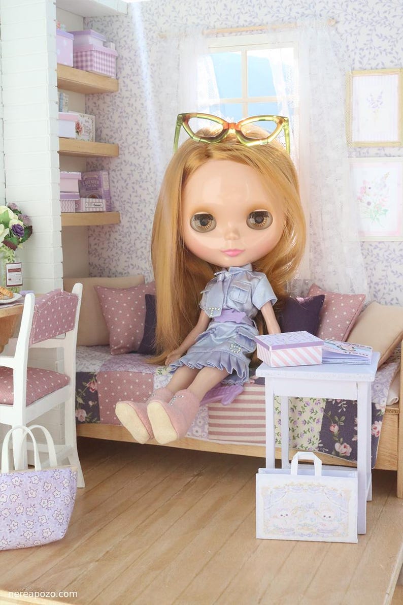 Pode incluir: Uma boneca com longos cabelos loiros e olhos grandes, vestida com um vestido azul claro e botas rosa, com &oacute;culos na cabe&ccedil;a. A boneca est&aacute; sentada em uma cama em miniatura com uma colcha de retalhos. Uma pequena mesa branca cont&eacute;m uma caixa e livros.