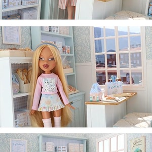 May include: A dollhouse bakery scene featuring a blonde doll in a pink long-sleeve shirt and patterned skirt, standing in a miniature shop with light blue shelves, white counters, and various tiny bakery items including boxes labeled Bakery.