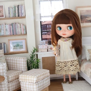 K&ouml;nnte beinhalten: Eine Puppe mit braunen Haaren und gro&szlig;en blauen Augen tr&auml;gt ein cremefarbenes Kleid mit Blumenmuster an den &Auml;rmeln und einem Spitzenbesatz. Die Puppe steht in einem Miniaturzimmer mit einem karierten Sessel, einem passenden Hocker und B&uuml;cherregalen.