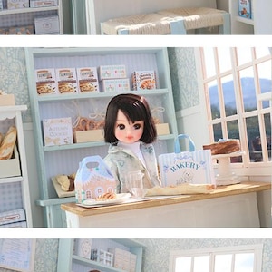 May include: A miniature bakery scene featuring a doll with dark hair, a light-colored jacket, and a white top. The bakery includes shelves stocked with boxes of cookies, bread, and pastries, a wooden counter, and a blue-and-white striped bakery bag.
