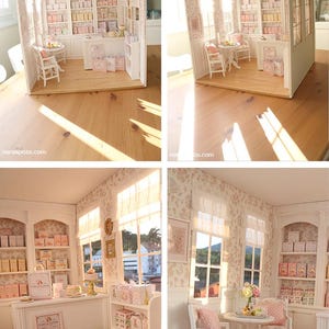 K&ouml;nnte beinhalten: Ein Miniatur-Puppenhaus-B&auml;ckereiladen mit wei&szlig;en Holzm&ouml;beln, einem runden Tisch mit zwei St&uuml;hlen, einer Verkaufstheke und Regalen voller kleiner Schachteln. Die W&auml;nde haben eine florale Tapete und der Laden enth&auml;lt Miniatur-Einkaufst&uuml;ten mit der Aufschrift Cake & Clover.