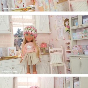 May include: A doll in a floral dress stands in a miniature kitchen scene. The doll has long brown hair and is surrounded by miniature furniture, including a cabinet, chair, and table. The scene is decorated in soft pink tones.