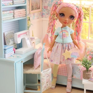K&ouml;nnte beinhalten: Eine Puppe in einem pastellfarbenen Zimmer mit Schreibtisch, B&uuml;cherregal und Bett. Die Puppe hat rosa und Regenbogen-Z&ouml;pfe, tr&auml;gt ein hellblaues Oberteil, einen rosa Rock und eine sternf&ouml;rmige Handtasche. Der Raum ist mit floraler Tapete und Miniaturm&ouml;beln dekoriert.