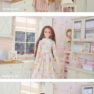 May include: A dollhouse kitchen scene with two dolls. One doll is in a striped dress, the other in a floral dress. The kitchen has white cabinets, a window, and decorative items, including books and a miniature chair.