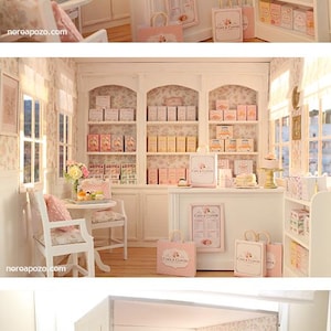K&ouml;nnte beinhalten: Ein Miniatur-Puppenhaus-B&auml;ckereiladen mit wei&szlig;en Regalen voller kleiner Schachteln, einer Verkaufstheke mit Einkaufst&uuml;ten mit der Aufschrift Cake & Clover, einem kleinen Tisch mit St&uuml;hlen, gebl&uuml;mter Tapete und Fenstern mit einfallendem Tageslicht.