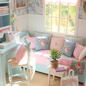 K&ouml;nnte beinhalten: Miniatur-Puppenhaus-Interieur in Pastellt&ouml;nen von Blau und Rosa. Der Raum verf&uuml;gt &uuml;ber ein kleines Sofa mit Blumendekokissen, eine N&auml;hmaschine und einen kleinen Tisch mit einer Topfpflanze. Die W&auml;nde sind mit gemusterter Tapete und einem gerahmten Bild geschm&uuml;ckt.