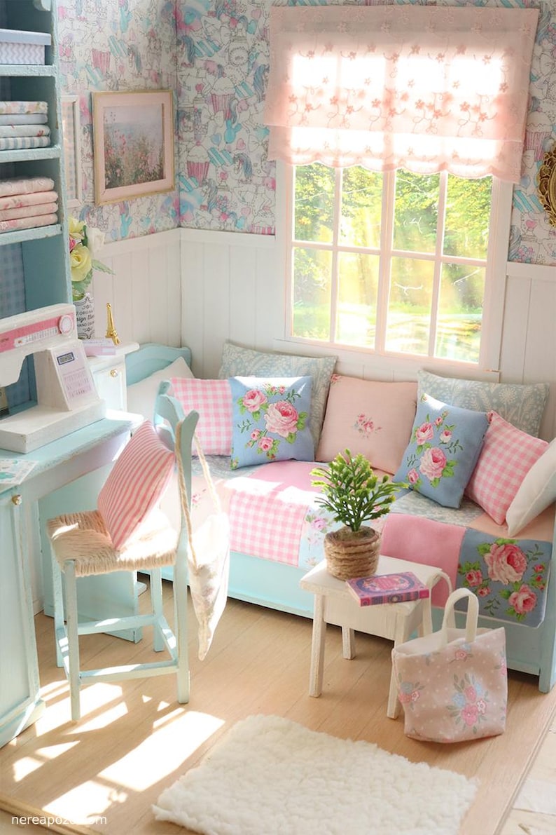 K&ouml;nnte beinhalten: Miniatur-Puppenhaus-Interieur in Pastellt&ouml;nen von Blau und Rosa. Der Raum verf&uuml;gt &uuml;ber ein kleines Sofa mit Blumendekokissen, eine N&auml;hmaschine und einen kleinen Tisch mit einer Topfpflanze. Die W&auml;nde sind mit gemusterter Tapete und einem gerahmten Bild geschm&uuml;ckt.