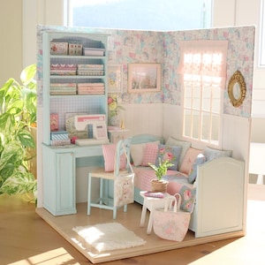 K&ouml;nnte beinhalten: Ein Miniatur-Puppenhaus-Zimmer mit einem hellblauen Schreibtisch, B&uuml;cherregal und Sofa. Der Raum ist mit floraler Tapete, rosa und blauen Kissen und einer kleinen N&auml;hmaschine dekoriert. Ein wei&szlig;er Teppich und eine Tragetasche liegen auf dem Boden.