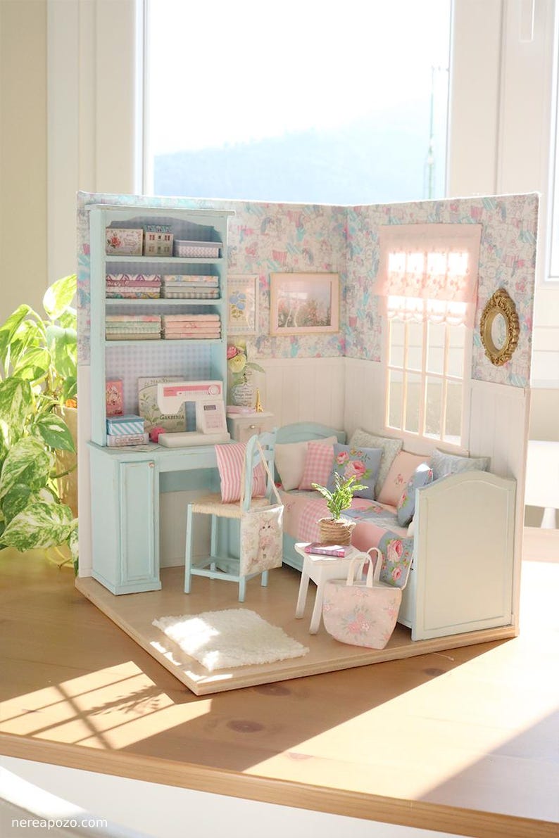 K&ouml;nnte beinhalten: Ein Miniatur-Puppenhaus-Zimmer mit einem hellblauen Schreibtisch, B&uuml;cherregal und Sofa. Der Raum ist mit floraler Tapete, rosa und blauen Kissen und einer kleinen N&auml;hmaschine dekoriert. Ein wei&szlig;er Teppich und eine Tragetasche liegen auf dem Boden.