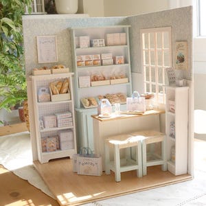 May include: A miniature bakery diorama featuring a light blue hutch, white shelving units, a wooden counter, and two stools. The shelves are stocked with tiny boxes, bread, and pastries. A window, wall art, and small paper shopping bags are included.
