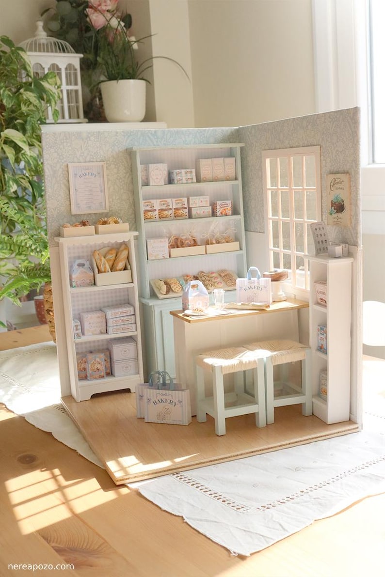 May include: A miniature bakery diorama featuring a light blue hutch, white shelving units, a wooden counter, and two stools. The shelves are stocked with tiny boxes, bread, and pastries. A window, wall art, and small paper shopping bags are included.