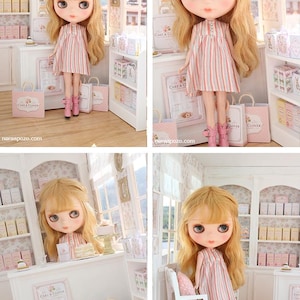 K&ouml;nnte beinhalten: Eine Puppe mit langem, welligem blonden Haar und gro&szlig;en Augen tr&auml;gt ein &auml;rmelloses Kleid mit vertikalen rosa, wei&szlig;en und blauen Streifen sowie rosa Stiefel. Die Puppe befindet sich in einer Miniatur-B&auml;ckerei mit Regalen und kleinen Einkaufst&uuml;ten.