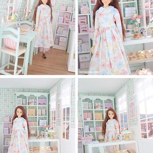 K&ouml;nnte beinhalten: Eine Puppe in einem Blumenkleid steht in einer Miniatur-B&auml;ckerei. Die B&auml;ckerei verf&uuml;gt &uuml;ber Regale mit verpackten Waren, eine Vitrine mit Geb&auml;ck und rosa B&auml;ckereitaschen. Die W&auml;nde sind mit einem hellgr&uuml;nen und wei&szlig;en Karomuster verziert.