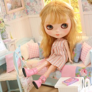 K&ouml;nnte beinhalten: Eine Blythe-Puppe mit blonden Haaren und gro&szlig;en Augen sitzt auf einem Miniatursofa mit rosa und blauen Blumenkissen. Die Puppe tr&auml;gt ein gestreiftes Kleid und rosa Stiefel. Ein kleiner Tisch und Stuhl sind in der N&auml;he.