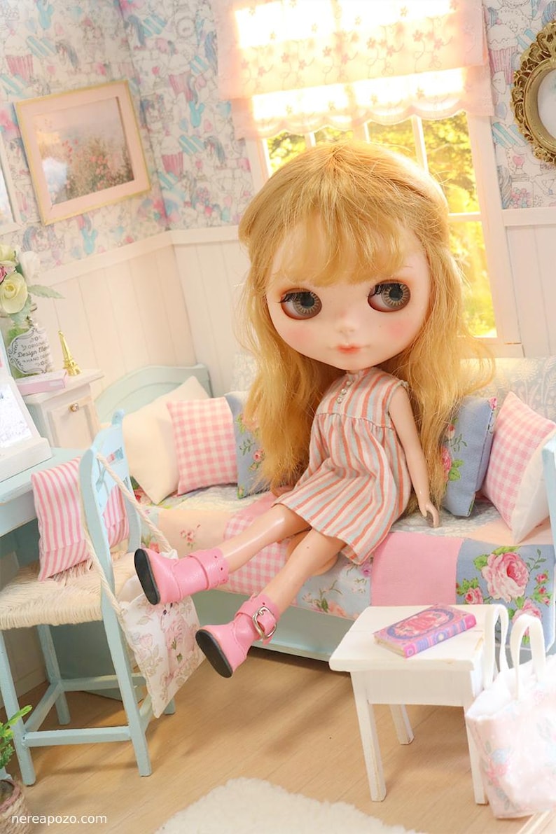 K&ouml;nnte beinhalten: Eine Blythe-Puppe mit blonden Haaren und gro&szlig;en Augen sitzt auf einem Miniatursofa mit rosa und blauen Blumenkissen. Die Puppe tr&auml;gt ein gestreiftes Kleid und rosa Stiefel. Ein kleiner Tisch und Stuhl sind in der N&auml;he.