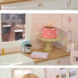 May include: Miniature kitchen scene with a doll, cookbooks, and baking supplies. Includes a pink cake, cookie boxes, and a rolling pin. The scene is set in a dollhouse with a window and white cabinets.