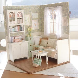 Może przedstawiać: Miniaturowy pok&oacute;j dla lalek z białymi meblami, w tym szafką, regałem i małym stolikiem. Pok&oacute;j ma kwiatową tapetę, okno z przezroczystymi zasłonami i dekoracyjne oprawione obrazy. Mała zielono-biała torba w kratkę leży na stole.