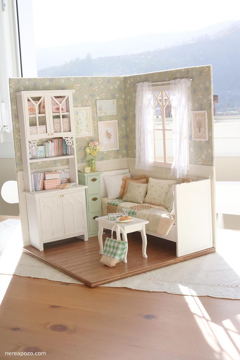 Może przedstawiać: Miniaturowy pok&oacute;j dla lalek z białymi meblami, w tym szafką, regałem i małym stolikiem. Pok&oacute;j ma kwiatową tapetę, okno z przezroczystymi zasłonami i dekoracyjne oprawione obrazy. Mała zielono-biała torba w kratkę leży na stole.