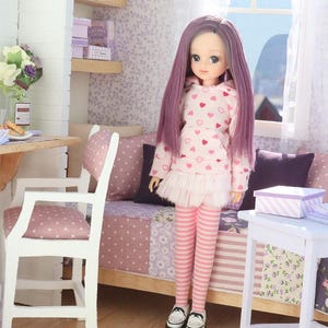 Pode incluir: Uma boneca com cabelo comprido roxo, vestindo um moletom rosa com padr&otilde;es de cora&ccedil;&atilde;o, uma saia branca e leggings listradas rosa. A boneca est&aacute; em um quarto em miniatura com uma cama, cadeira e prateleiras.
