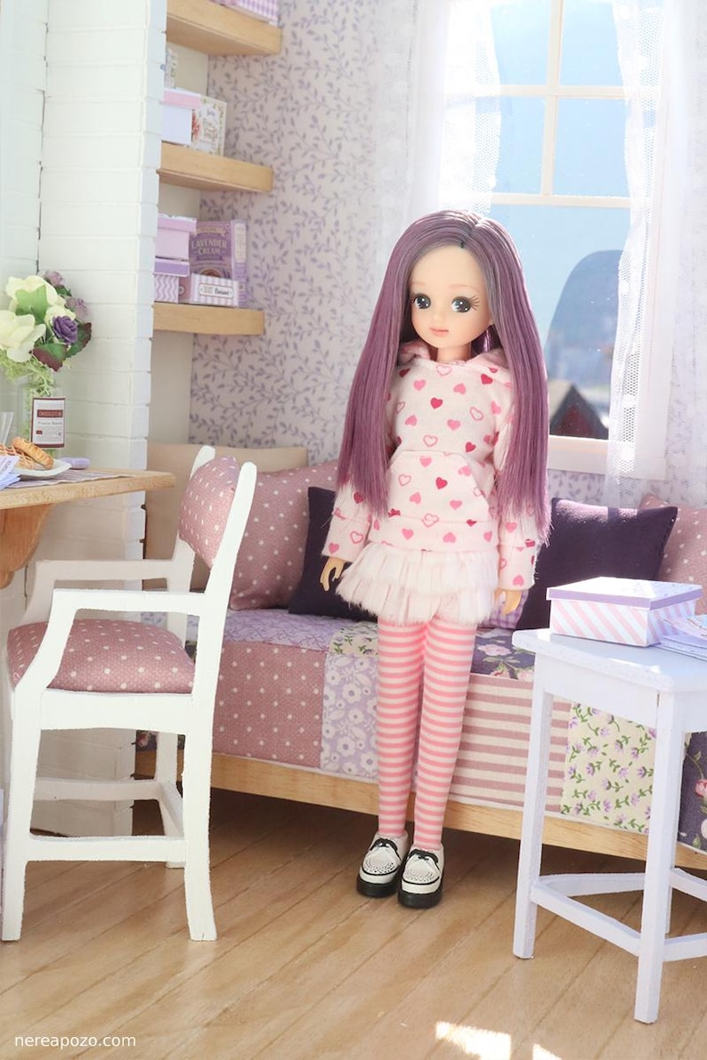 Pode incluir: Uma boneca com cabelo comprido roxo, vestindo um moletom rosa com padr&otilde;es de cora&ccedil;&atilde;o, uma saia branca e leggings listradas rosa. A boneca est&aacute; em um quarto em miniatura com uma cama, cadeira e prateleiras.