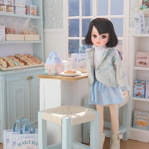 May include: A doll with dark hair wearing a light blue floral jacket, white turtleneck, and light blue skirt with white boots, standing in a miniature bakery shop with shelves of food, a small table, a stool, and bakery-themed paper bags.