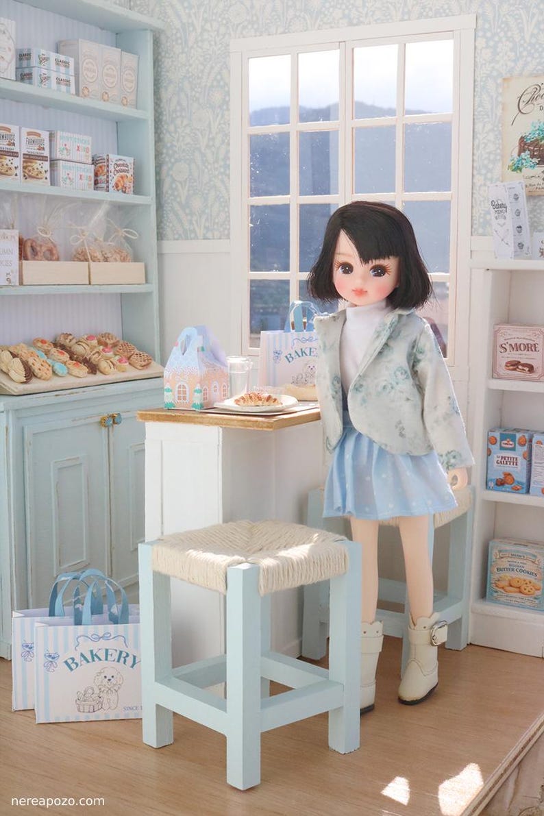 May include: A doll with dark hair wearing a light blue floral jacket, white turtleneck, and light blue skirt with white boots, standing in a miniature bakery shop with shelves of food, a small table, a stool, and bakery-themed paper bags.