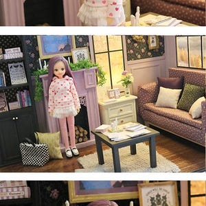 May include: A doll with long purple hair wearing a white hooded sweatshirt featuring a pink heart pattern, a white pleated skirt, and pink and white striped leggings. The doll has large brown eyes and is standing in a miniature room setting.