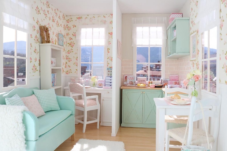 May include: A detailed dollhouse interior with a living room and kitchen. The living room has a teal sofa with cushions, a white bookshelf, and floral wallpaper. The kitchen features a mint green cabinet and a white table.