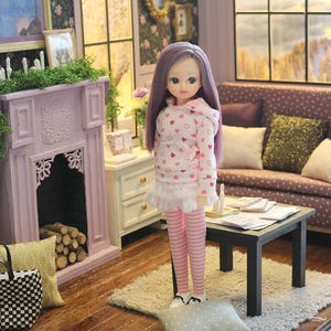 May include: A doll with long purple hair wearing a white hooded sweatshirt with a pink heart pattern, a white ruffled skirt, and pink and white striped leggings. The doll stands in a miniature room setting with a fireplace, sofa, and table.