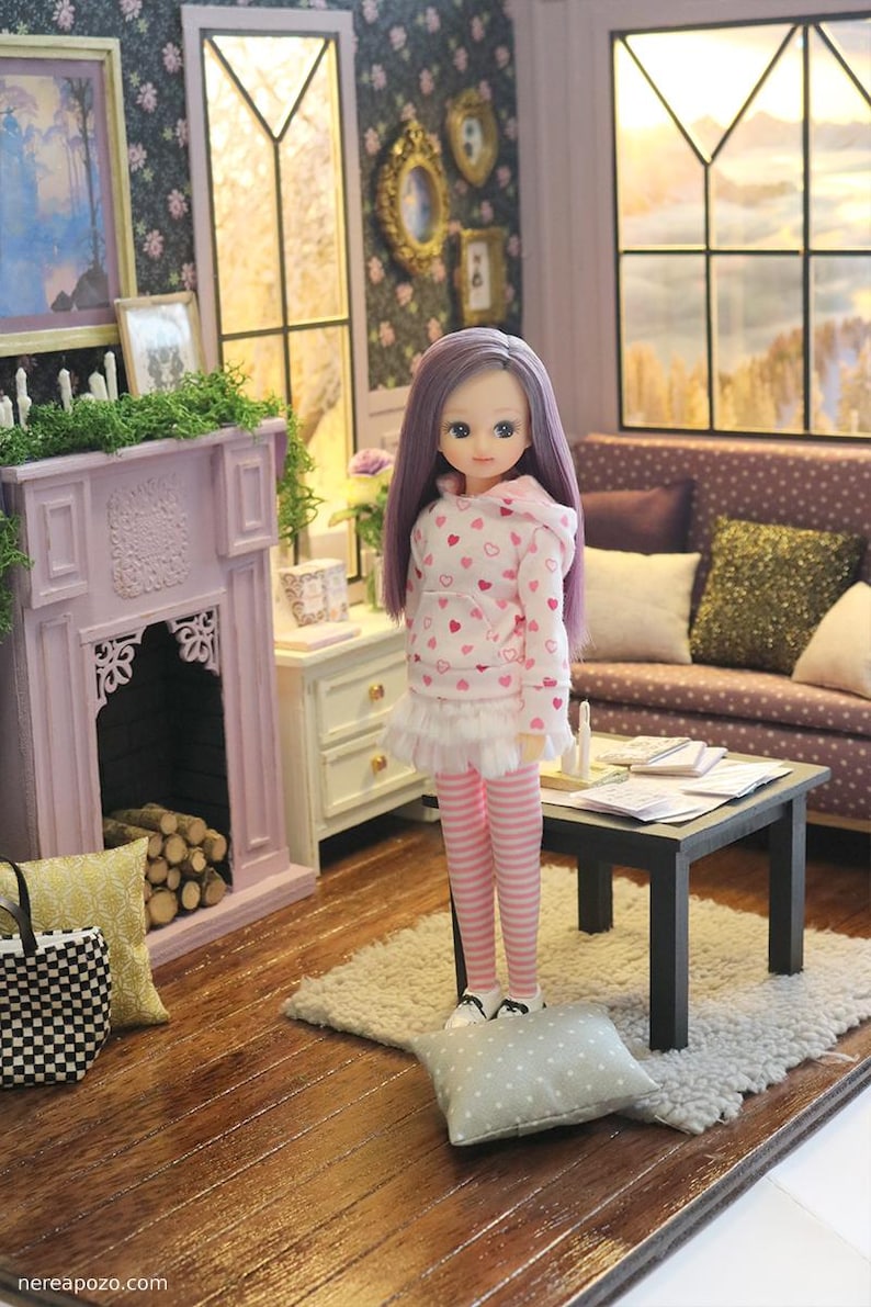 May include: A doll with long purple hair wearing a white hooded sweatshirt with a pink heart pattern, a white ruffled skirt, and pink and white striped leggings. The doll stands in a miniature room setting with a fireplace, sofa, and table.