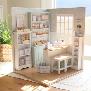 Könnte beinhalten: Ein Miniatur-Bäckerei-Diorama mit hellblauen und weißen Holzregalen, gefüllt mit winzigem Brot, Gebäck und Schachteln. Die Szene umfasst eine kleine Theke mit zwei Hockern, Papiereinkaufstüten mit der Aufschrift BAKERY und Wandbilder.